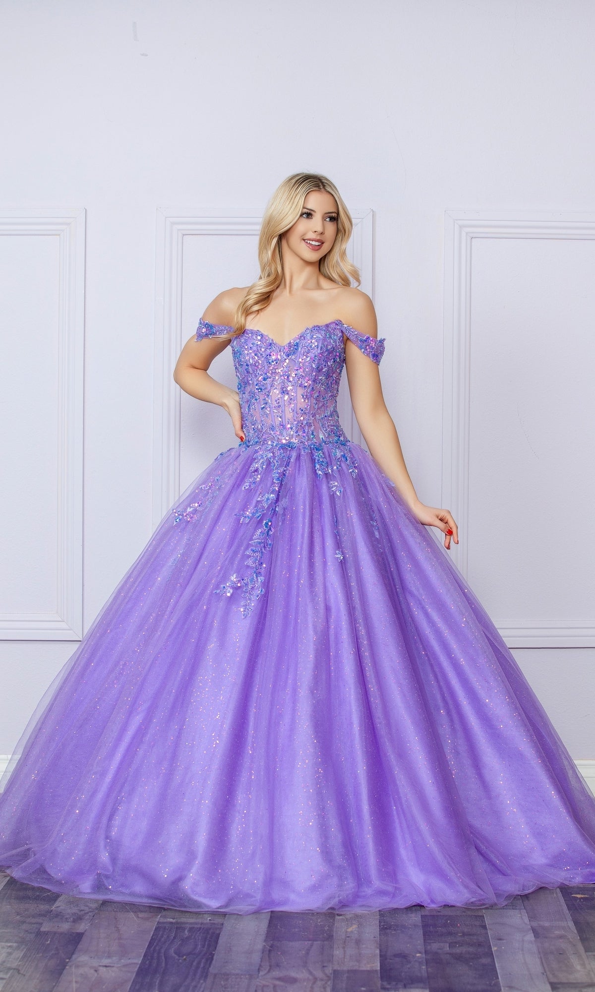 A woman wearing the Bright Off-the-Shoulder Long Prom Ball Gown H1349, featuring a sparkly design and full tulle skirt, stands in front of a white paneled wall.